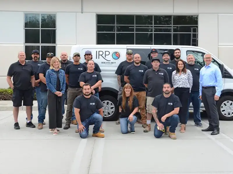 Group of 18 people posing in front of a white van with IRP logo outside a building.