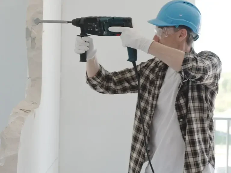 Worker in a blue helmet using a power drill to break a wall indoors.