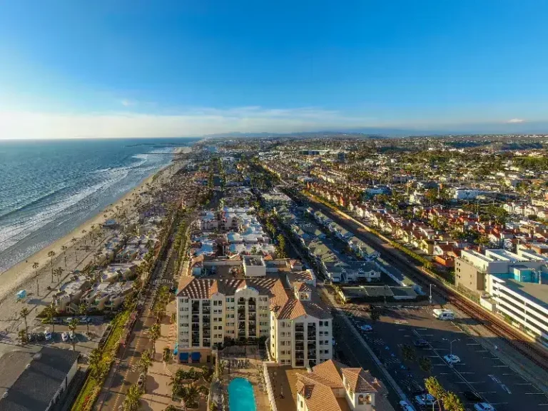 Coastal cityscape with beachfront, residential buildings, and a swimming pool under clear blue sky.