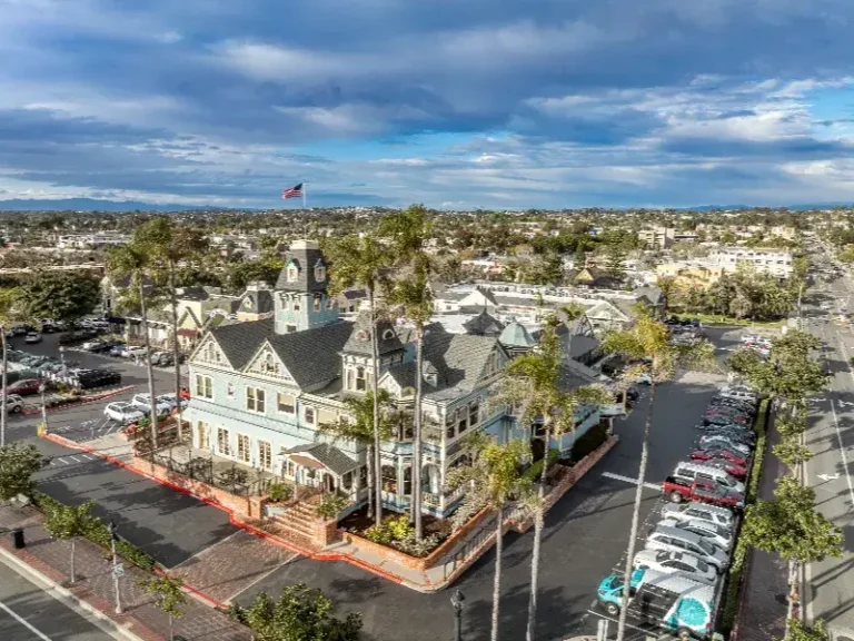 Large Victorian-style building surrounded by palm trees and parking lots under a cloudy sky.