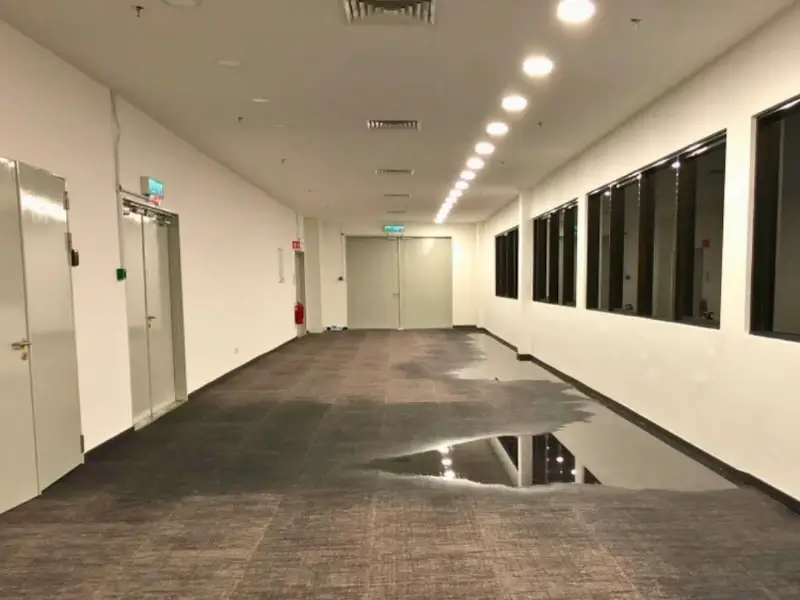 Empty hallway with carpeted floor and a large puddle of water reflecting ceiling lights.