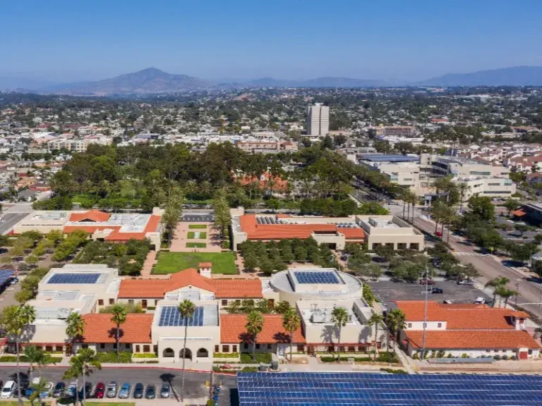 Aerial view of a large building complex with red roofs and solar panels, surrounded by trees and cityscape.