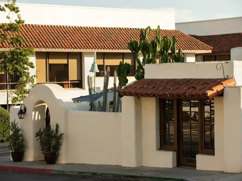 White stucco building with terracotta roof tiles and tall cacti behind a low wall.