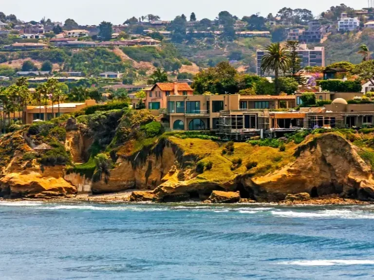 Coastal houses atop rocky cliffs overlooking the ocean with hills and trees in the background.