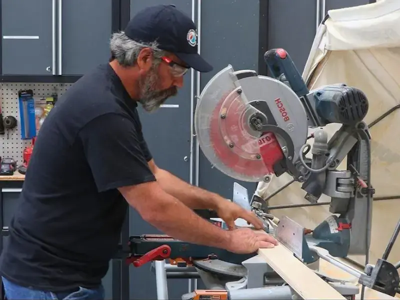 Man wearing safety glasses and a cap uses a miter saw to cut a wooden board.
