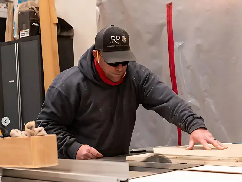 Man wearing a cap and hoodie cutting wood on a table saw in a workshop.