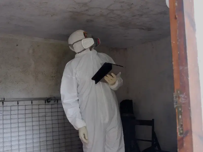 Person in protective suit and mask inspecting a ceiling while holding a tablet inside a dim room.
