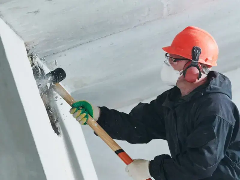 Worker in safety gear breaking concrete wall with sledgehammer.