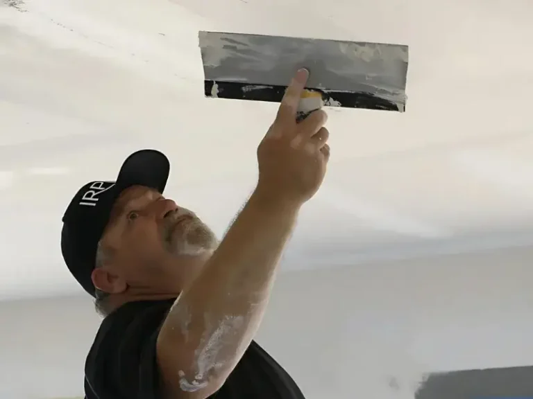 Man wearing a cap applying plaster or joint compound to a ceiling with a drywall taping knife.