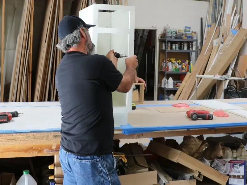 Man using a power drill on a white wooden cabinet in a workshop.