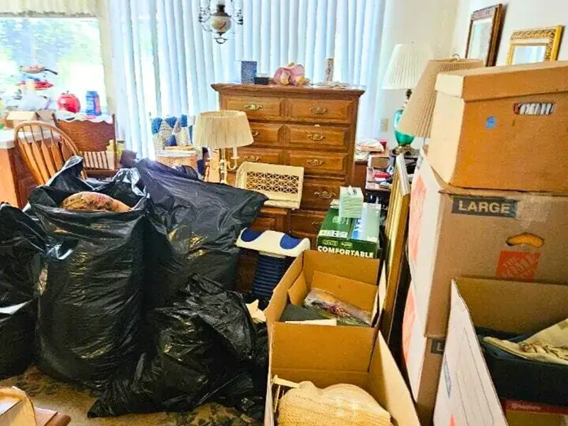 Packed moving boxes and black trash bags clutter a living room with wooden furniture and lamps.