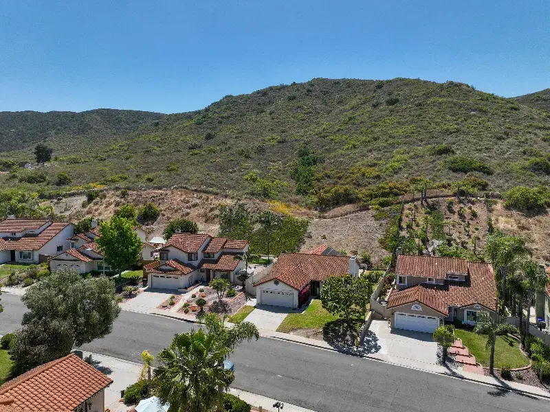 Suburban houses with red-tiled roofs along a street, backed by green hills under a clear blue sky.