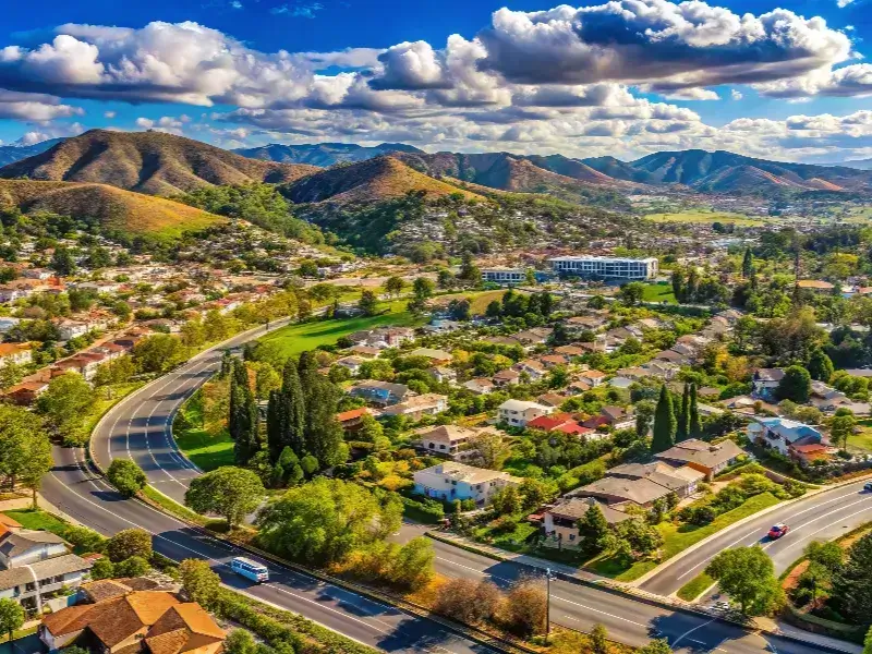 Suburban neighborhood with winding roads, houses, and hills under a partly cloudy sky.